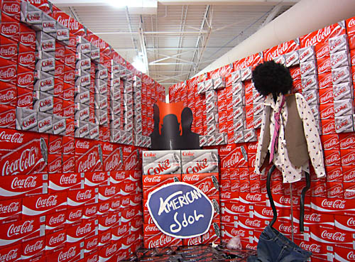 Giant display of Coke cases spells out IDOL on both walls, has a 
makeshift ‘American Idol’ logotype, and has a hanged effigy with its 
pants down