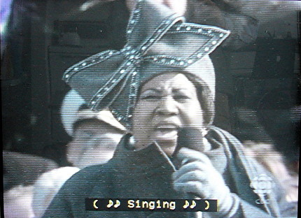 Aretha Franklin wears a grey outfit with matching hat and huge bow. Caption reads ( ♪♪ Singing ♪♪ ) 