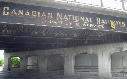 Railway overpass above a road reads CANADIAN NATIONAL RAILWAY COURTESY & SERVICE in distressed white letters
