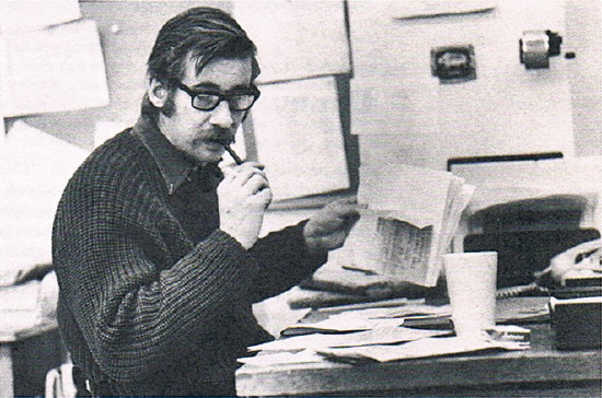 Peter Gzowski, in heavy cabled sweater, ruffles papers and looks at desk while smoking a pipe