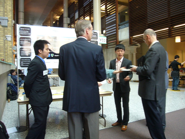 Two students and two old guys in suits chat in front of the students’ poster