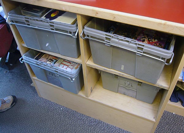 Four grey bins under a counter, all full of books