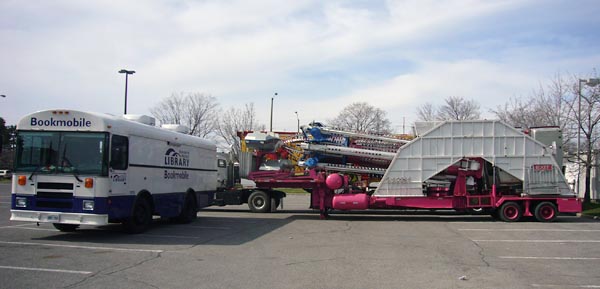 Bookmobile parked alongside and pointing away from a set of folded silver-and-blue booms on a hot-pink trailer