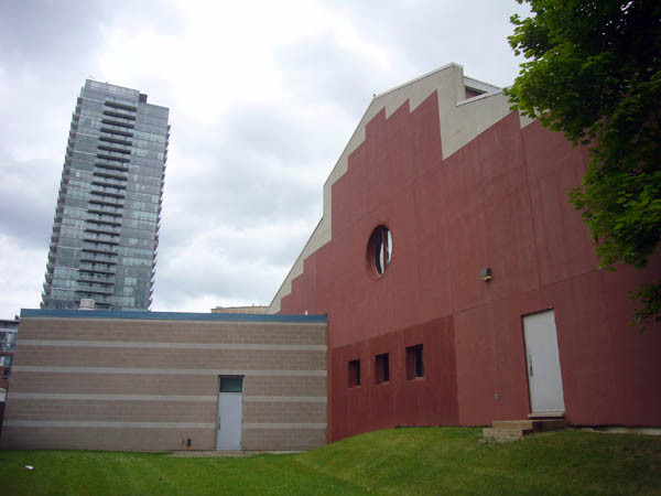 Barn-like end of building is clad in red brick with a white border and central porthole window. Alongside, a one-story beige addition. In the distance, a condo tower