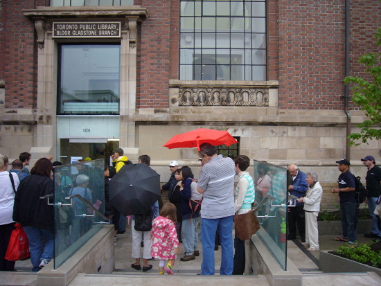 People, many carrying umbrellas, wait outside a brick building with an old sign reading TORONTO PUBLIC LIBRARY BLOOR GLADSTONE BRANCH