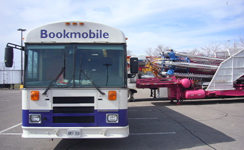 Front of Bookmobile bus next to the profile of large industrial apparatus folded onto a pink flatbed trailer