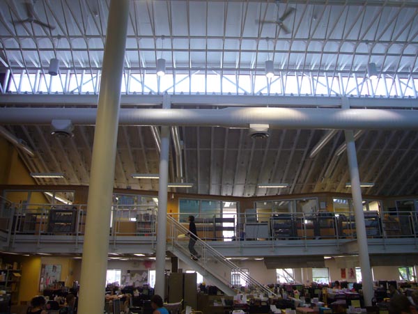 Mezzanine below a ceiling with side-facing skylights, white pipes that look like ribs, and ceiling fans