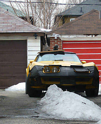 Yellow Corvette with black bra over front end parked between two houses
