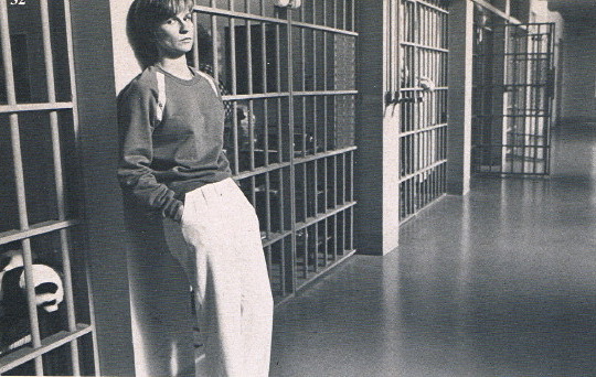 Young woman in sweatshirt and white trousers leans against cellblock doors in prison hallway