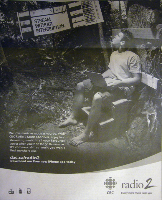 Shoeless young man in T-shirt and shorts sits with laptop in dense forest