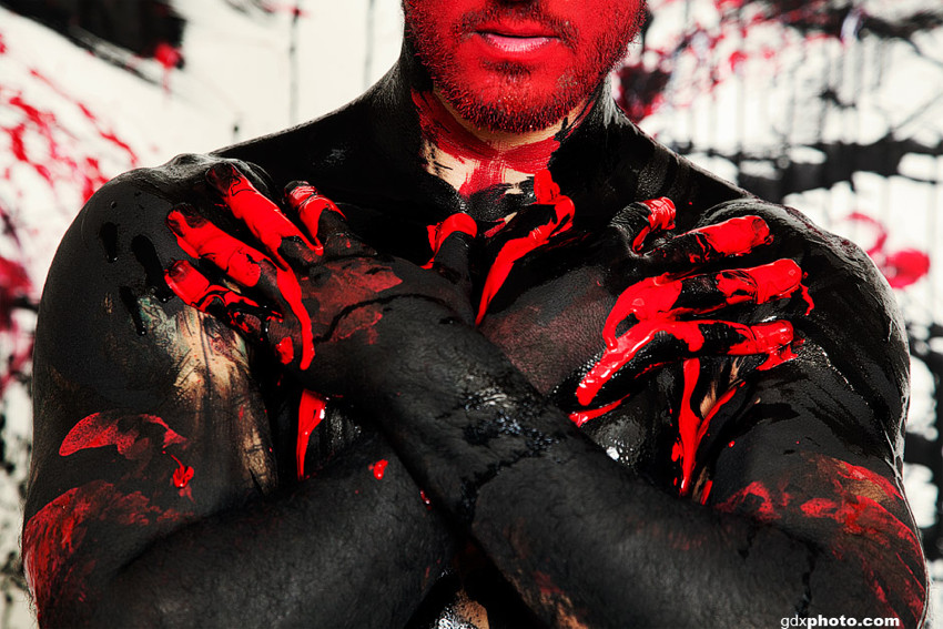 Shirtless man in black bodypaint has red-painted face, red-paint-smeared fingers crossed over chest