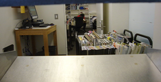 Carts of books and DVDs, a computer on a bench, and a distant staffmember seen through a blue and silver slot