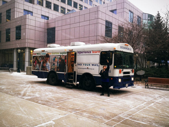 Woman adjusts right-side mirror of bookmobile, parked on snowy concrete slabs