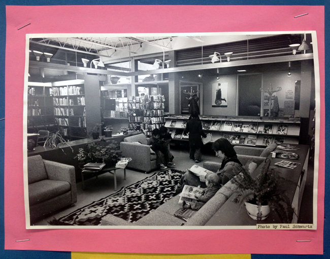 Black-and-white photo shows living-room-like couch, armchairs, end table under track lighting