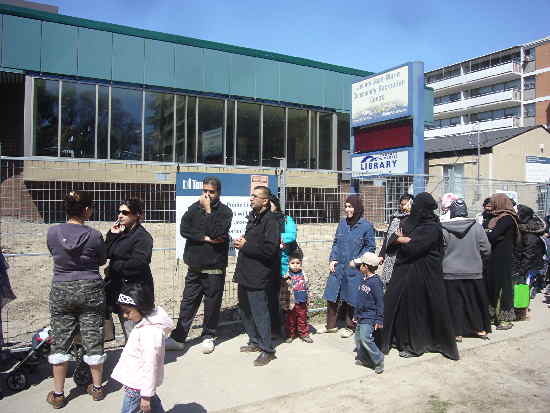 3 kids, 2 men, 5 women not in hijab or niqab, 4 in hijab, 1 in niqab, 1 undetermined Queue of people outside community centre and library