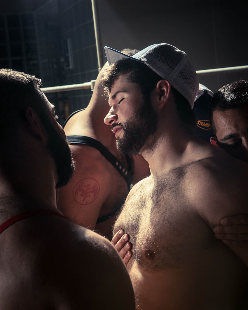 Young man, eyes shut and in a trucker cap, stands in a shaft of light in a throng of other men, one gazing at him, his own fingers pressed against his chest