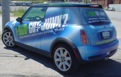 Rear three-quarter view of a blue Mini Cooper emblazoned with ‘1-800-GOT-JUNK?’ and other slogans