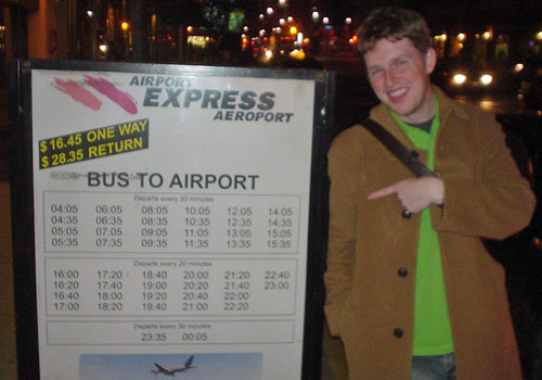 Matt Müllenweg, in green shirt and brown jacket, points jauntily at a sign for AIRPORT EXPRESS AÉROPORT giving BUS TO AIRPORT times in Arial