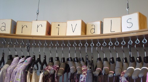 Wooden blocks atop shelf that supports a store’s clothing racks read ‘arrivals’ in hand-drawn Letter Gothic