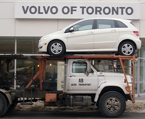 White B-class Mercedes on upper deck of auto carrier parked outside Volvo dealership