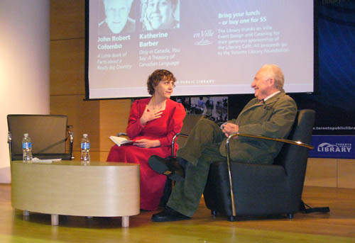 Barber and Colombo seated onstage in front of Toronto Public Library slide showing their names and faces