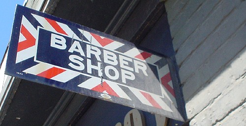 Sign projecting from wall has red, white, and blue X-shaped stripes and BARBER SHOP in white sansserif on a blue background