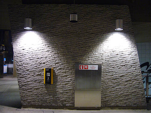 Two spotlights illuminate a dark wall of rough-surfaced bricks mounted at an angle