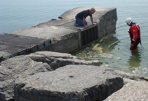 Man in street clothes crouches over intake vent on rocky outcropping in lake while another man in orange wetsuit and white helmet walks toward the vent