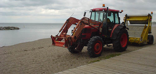 On a sandy beach between a boardwalk and the water, a red tractor pulls yellow trailer whose front face consists of steel cylinders