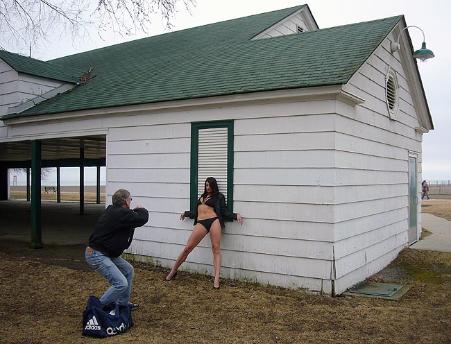 Black-haired woman in black bikini and jacket splays legs and leans against building as a middle-aged guy in jeans snaps a picture