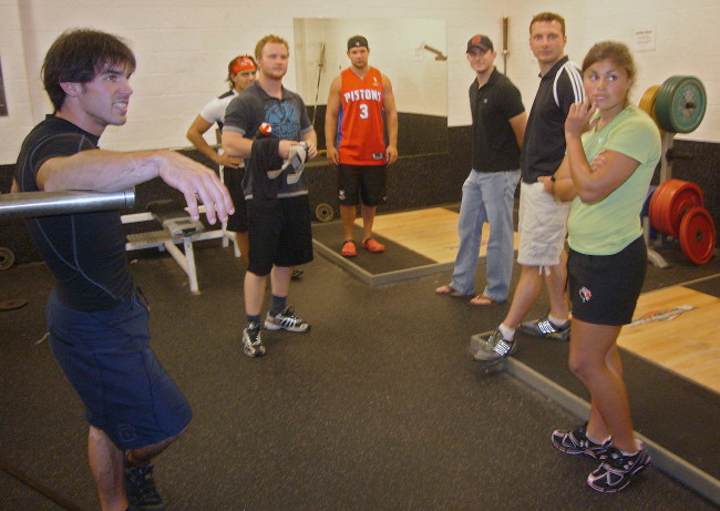 Five athletes in weightroom, along with Florian and James, turn toward right of camera