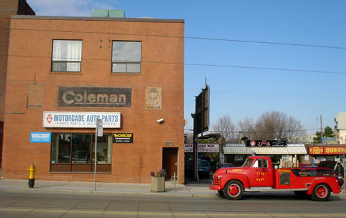Vintage red fire engine sits parked just down the street from a brick building with faded Coleman sign and new Motorcade Auto Parts sign