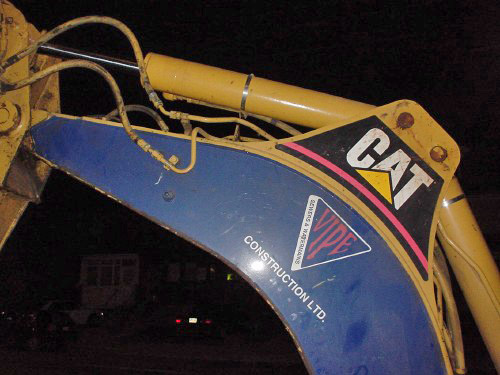 Nighttime shot shows the crown of a yellow backhoe’s arm emblazoned with ‘CAT.’ The blue-painted articulated arm reads ‘Vipe Construction Ltd.’
