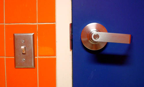 Bright orange tiled wall, with metal-faced light switch, sits next to a white door jamb and a deep blue door with silver metal handle