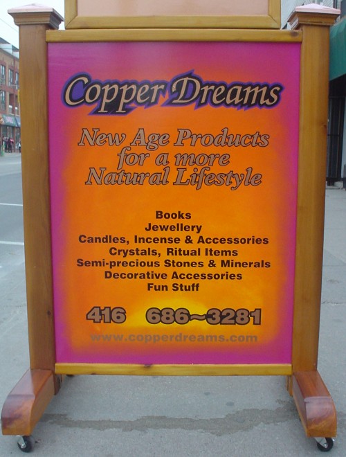 Pink, gold, and yellow sandwich-board sign with script and italic type: ‘Copper Dreams: New Age Products for a more Natural Lifestyle’