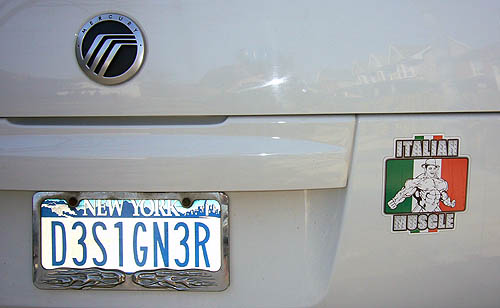 New York state vanity licence plate on a Mercury SUV reads D3S1GN3R. Nearby, a green-white-red ITALIAN MUSCLE bumper sticker shows a shirtless bodybuilder in a hard hat