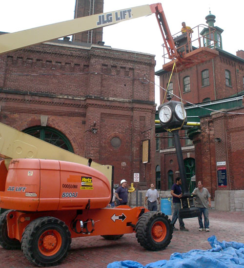 Men steady a tall black four-sided clock as a crane lowers it into position on a cobblestone street