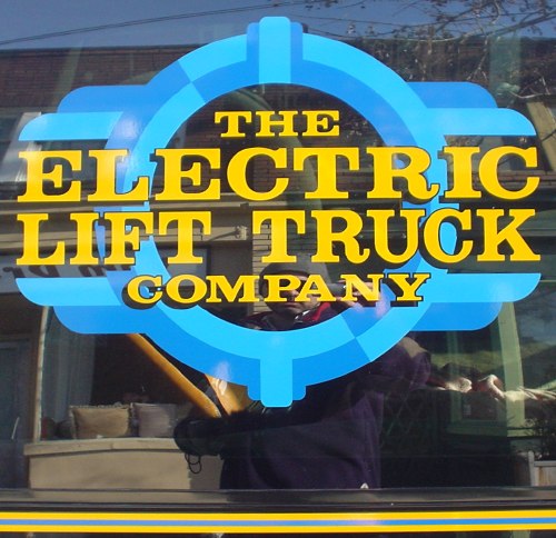 Painted window sign with yellow type reading ‘The Electric Lift Truck Company’ on a bright-blue shield