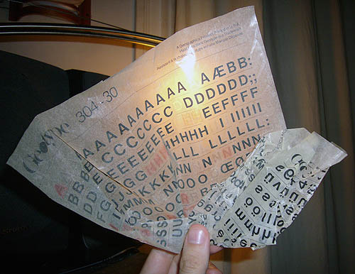 Fingers hold crinkled sheet of Geotype instant lettering (Helvetica typeface) in front of a light