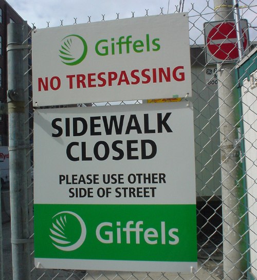 Two warning signs outside construction site, with black, green, and red Frutiger type