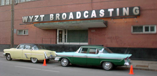 Brick building has sign over entrance reading WZYT BROADCASTING in Arial Black, with two vintage cars parked on the street next to orange pylons