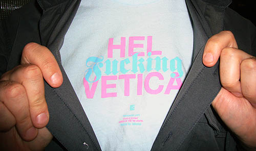 Hands hold open a shirt to reveal a blue T-shirt with pink-and-blue type reading HEL Fucking VETICA