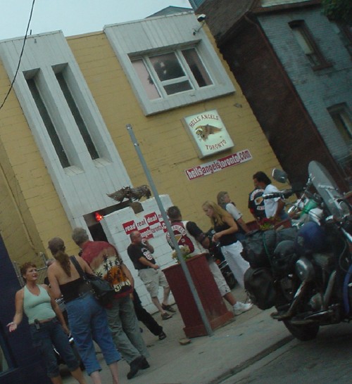 Men in T-shirts and vests and women in tank tops and tube tops mill about outside building labeled Hells Angels Toronto