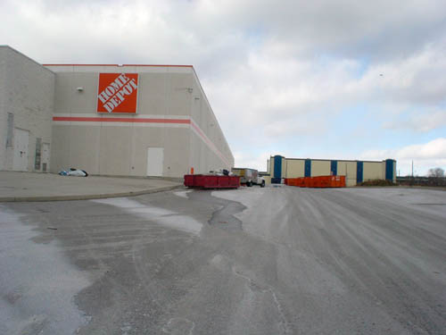 Barren greay-and-white parking ramp leads to barren grey-and-white two-storey concrete building with orange Home Depot sign and single orange stripe. Warehouse and dumpsters sit on the ramp