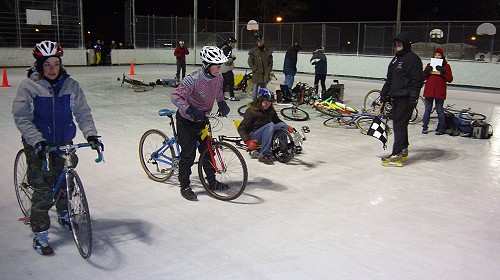 Two riders on stand-up bikes and a third on a recumbent wait alongside a man holding a starting flag