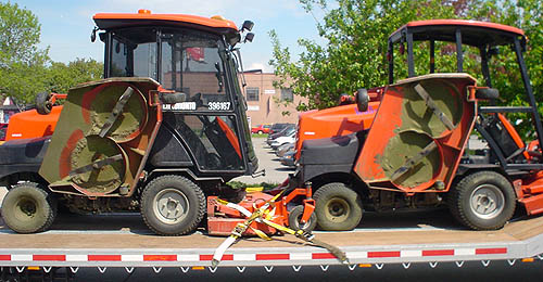 Two red, tractor-sized industrial lawnmowers sit with blades pulled upright on a flatbed trailer
