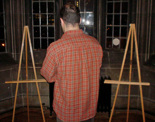 Balding man in red plaid shirt faces away and stands in a bay window between empty wooden easels