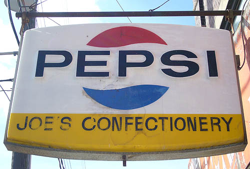 Sign with the word PEPSI between red and blue quarter-circle waves above and below, and JOE’S CONFECTIONERY on a yellow background