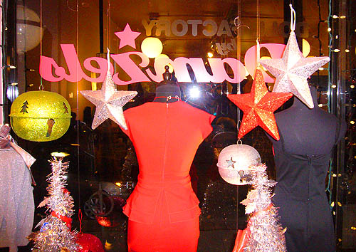Brightly-coloured shop vitrine, seen from behind, has orange women’s suit, garlands, hanging ornaments, and Damzel’s written on the glass