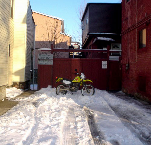 Yellow motorcycle sits in profile at end of snow-covered driveway between two houses